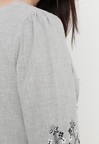 Grå linnetopp med puffärmar, prydd med blommig brodyr i svart och vitt på manschetterna. Slät textur med en avslappnad design.