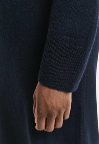 Navy woolen sweater detail showcasing a ribbed cuff and subtle textured pattern, with a visible logo on the sleeve.