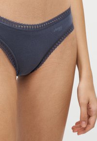 Buste féminin portant un sous-vêtement style bikini bleu marine avec bordure en dentelle, cuisses supérieures visibles et main droite partielle sur fond uni.