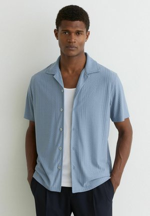 Mężczyzna stojący przy białej ścianie, ubrany w jasnoniebieską, krótkorękawową koszulę na guziki noszoną na białym podkoszulku oraz ciemne spodnie, z rękami w kieszeniach.