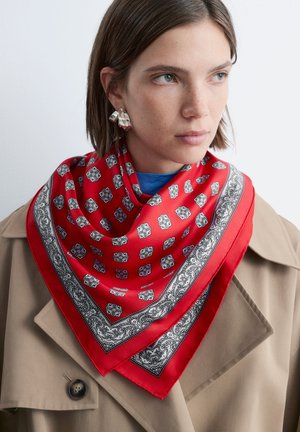 Jonge vrouw met een beige jas en een rood zijden sjaaltje met patroon om haar nek, met kort bruin haar en zilveren oorbellen.