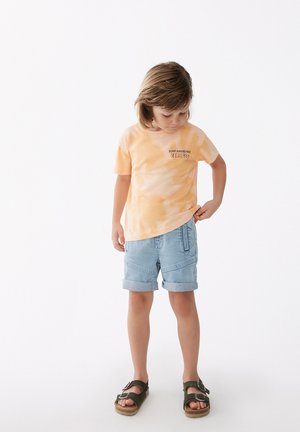 Camiseta de tie-dye naranja con mangas cortas, que presenta un texto, combinada con pantalones cortos de mezclilla claros y sandalias marrones con tirantes ajustables.