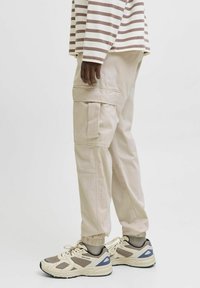 Beige Cargohosen mit elastischen Bündchen und mehreren Taschen, kombiniert mit hellfarbenen Sneakers mit Mesh- und Wildlederakzenten. Gestreiftes Oberteil sichtbar.