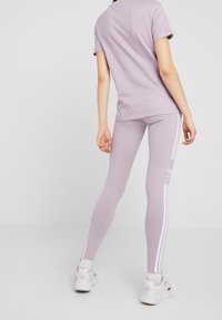 Fialové bavlněné tričko s krátkými rukávy spárované s odpovídajícími legínami se bílými bočními pruhy a logem, doplněné bílými teniskami.