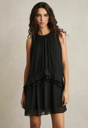Mujer con cabello oscuro y rizado, que lleva un vestido negro sin mangas con detalles de volantes en capas y dobladillo transparente, de pie sobre un fondo neutro.