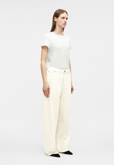 Femme aux cheveux bruns raides portant un t-shirt blanc ajusté, un pantalon large écru et des chaussures noires pointues, debout devant un fond uni.