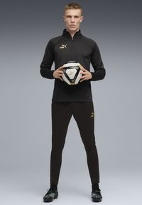 Svarta sportkläder med guldiga detaljer, inklusive en zip-up tröja och åtsittande byxor. Svart och vit fotboll hålls framför.