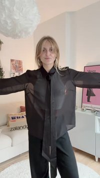 Blusa preta transparente com gola amarrada, mangas longas e fecho de botões. O tecido é leve, criando uma aparência translúcida.