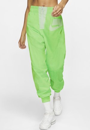 Femme portant un pantalon de jogging Nike vert vif avec une taille élastique, une fermeture éclair argentée, des chaussettes blanches et des baskets blanches, marchant devant un fond uni.