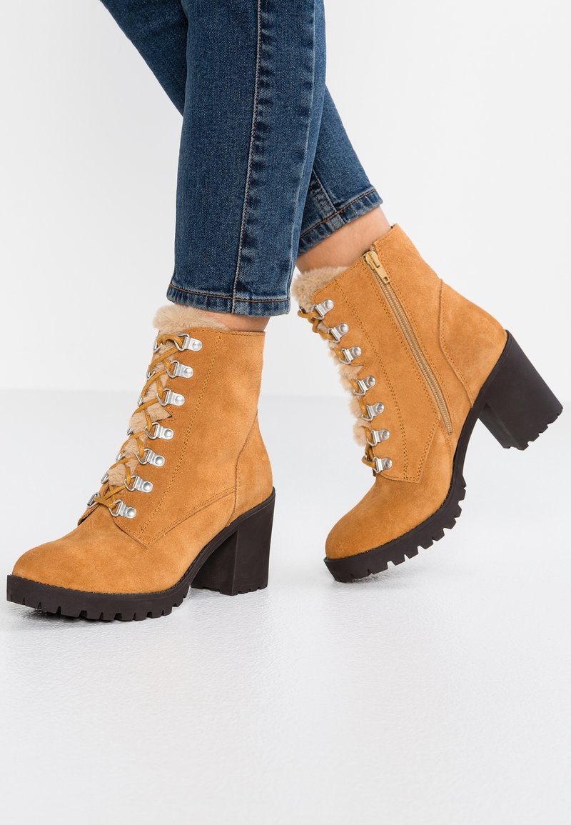 Bottines en daim fauve avec fermeture à lacets sur le devant, doublure en fausse fourrure, fermeture éclair latérale, et talon épais en caoutchouc noir. Semelle texturée pour une meilleure adhérence.