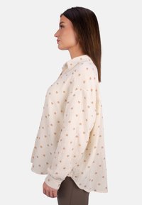 Cremfarbige Bluse mit Knopfleiste aus strukturiertem Stoff, verziert mit kleinen braunen Herzmustern. Sie hat einen lockeren Schnitt und lange Ärmel.