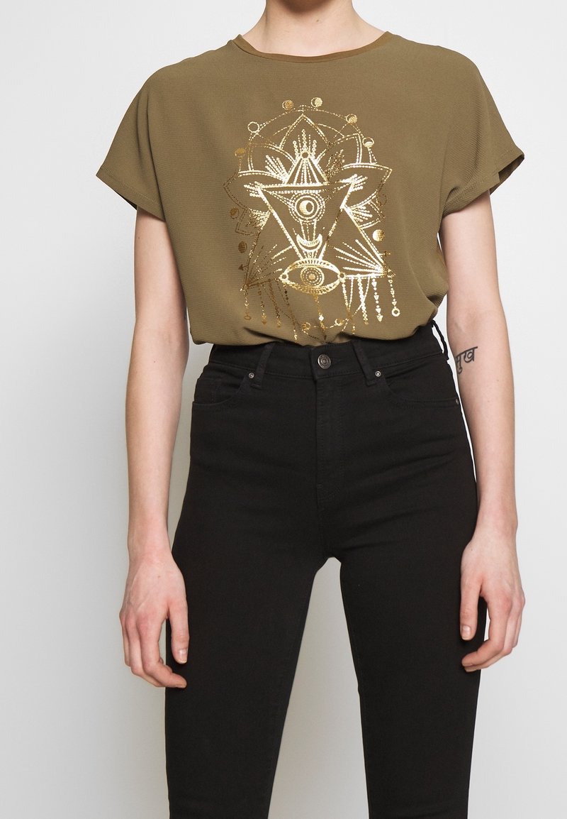 T-shirt à manches courtes vert olive avec un motif géométrique doré. Associé à un jean noir ajusté avec fermeture à boutons sur le devant et poches.