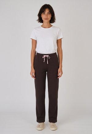 Mujer de pie con una camisa blanca de manga corta, pantalones marrón oscuro con cordón rosa y zapatillas beige contra un fondo claro y liso.