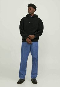 Giovane uomo in felpa nera con la scritta "Gymnas Studio", jeans blu e scarpe nere, in piedi contro uno sfondo chiaro, con lo sguardo rivolto di lato.
