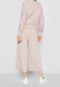 Donna che indossa una giacca bomber rosa chiaro, pantaloni beige a gamba larga e scarpe da ginnastica bianche, in piedi davanti a uno sfondo bianco semplice.