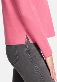 Różowy sweter wełniany z długimi rękawami, bocznymi rozcięciami i luźnym krojem, zestawiony z ciemnoszarymi spodniami typu slim fit. Widoczna gładka tekstura.