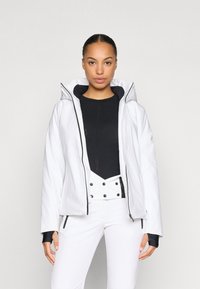 White jacket with a fitted design, hood, and black accents. Worn over a black top and white pants, showcasing a sleek, contemporary look.