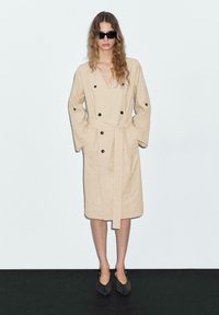 Beige Trenchcoat mit doppelreihigem Design, schwarzen Knöpfen, tailliertem Gürtel und weiten Ärmeln, kombiniert mit dunklen Sonnenbrillen und spitzen Ballerinas.