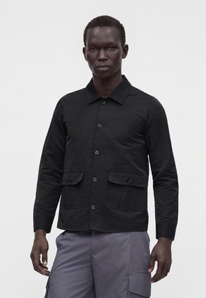 Hombre con una chaqueta negra abotonada con dos bolsillos frontales con solapa y pantalones cargo grises, de pie frente a un fondo liso y claro.