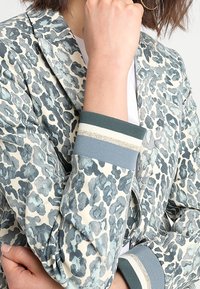 Blazer de estampado de leopardo con base de crema clara, que presenta manchas azules y grises, puños acanalados con rayas blancas y plateadas, y solapa de muesca.