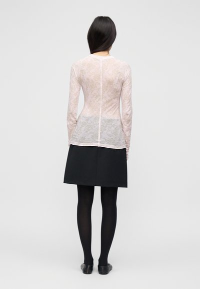 Svetlo roza prosojna majica z dolgimi rokavi in cvetličnim čipkastim vzorcem, kombinirana s črno krilo, teksturiranimi nogavicami in črnimi čevlji, videna od zadaj.