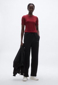 Piros rövid ujjú, testhezálló ing logóval, fekete bővebb szabású melegítőnadrág, fehér tornacipő, kezében fekete bombázó dzseki.