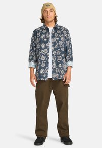 Bloemmotiefshirt in donkerblauw en lichtcrème, met lange mouwen en opgerolde manchetten. Gecombineerd met olijfgroene broek en zwarte schoenen.
