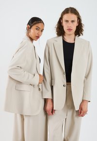 Blazers beige ajustés avec des revers, un bouton unique et des poches avant, associés à des tops noirs ajustés et à des pantalons beige assortis.