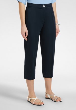 Frau trägt schwarze, knöchellange Hose, hellblaues Hemd, silbernes Armband und weiße flache Sandalen mit silbernen Verzierungen.