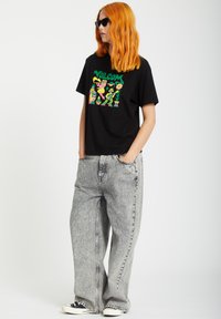 Zwarte graphic t-shirt met kleurrijk ontwerp, gecombineerd met hoogwater, lichtgrijze, loszittende jeans en zwarte sneakers met witte zolen.