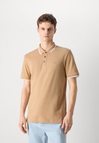 Bež polo majica s klasičnim ovratnikom, tri gumba na zapenjanju in belim robom na rokavih. Ima subtilen logotip blagovne znamke na prsih.