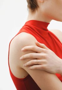 Anillo de oro con una pequeña piedra central, expuesto sobre una mano que descansa en un hombro desnudo. La figura lleva una blusa roja de cuello alto.