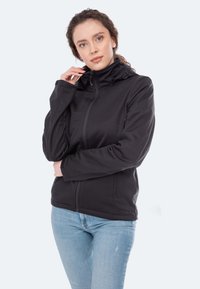 Schwarze Softshell-Jacke mit hohem Kragen, Frontreißverschluss und zwei Seitentaschen. Wird über blauen Jeans getragen und zeigt ein figurbetontes Design.
