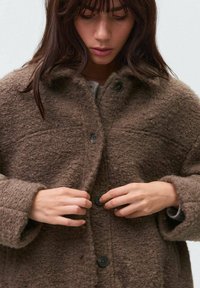 Palton texturat maro cu nasturi mari, croială lejeră și mâneci largi. Materialul pare moale și pufos, potrivit pentru vreme mai rece.