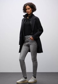 Abrigo negro con cuello alto, camiseta negra, jeans gris ajustados y zapatillas blancas. Presenta texturas suaves y líneas limpias.