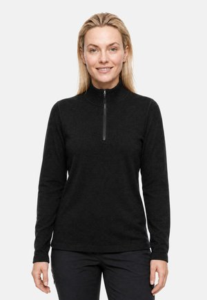 Femme souriante aux cheveux blonds portant un pull noir à manches longues avec une fermeture éclair devant, debout devant un fond clair uni.