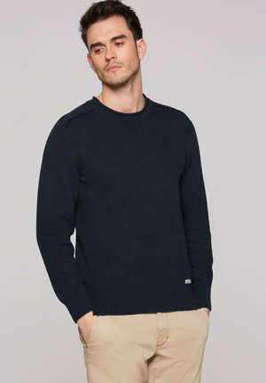Man wearing a dark blue crew neck sweater and beige pants, standing with hands in pockets, looking down and to the side against a gray background.