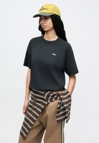 Schwarzes Baumwoll-T-Shirt mit kurzen Ärmeln und kleinem weißem "obey"-Logo, kombiniert mit brauner Hose und gelber Mütze mit grünen Akzenten.