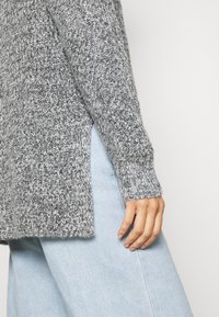 Gros plan d'une personne portant un pull en tricot gris avec une fente latérale et un jean en denim bleu clair, main reposant près de la hanche sur un fond blanc.