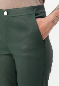 Mörkgröna skräddarsydda byxor i ett slätt material, med sidofickor, framknäppning och en mjuk yta.