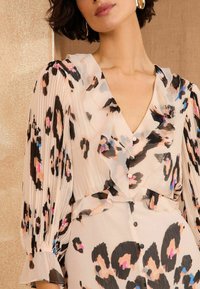 Pleated blouse pehmeas beežis, millel on leopardimuster musta, roosa ja sinise värviga. Omab V-dekolteed, nööpidega kinnitust ja volditud detaile.