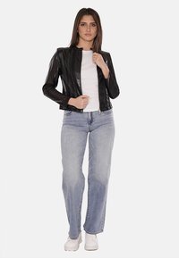 Giacca in pelle nera con chiusura a zip, design aderente, texture liscia e colletto rotondo, abbinata a jeans azzurri chiari e sneakers bianche.