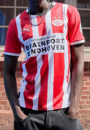 Personne portant un maillot de football rayé rouge et blanc du PSV Eindhoven avec texte du sponsor et logo Puma, mains dans les poches devant un mur en briques.