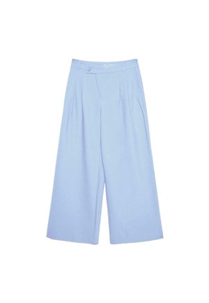 Personne portant un pantalon bleu clair taille haute avec des plis et une fermeture à boutons, mains dans les poches, associé à un top blanc court.
