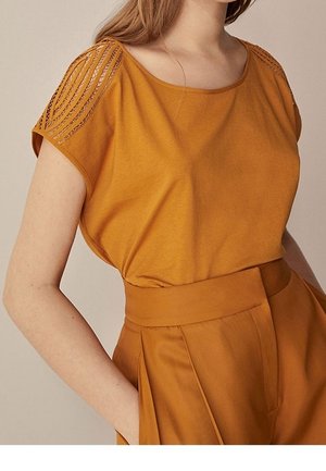 Mustard yellow top with a relaxed fit, featuring decorative lace detailing on the shoulders. Smooth texture and short sleeves.