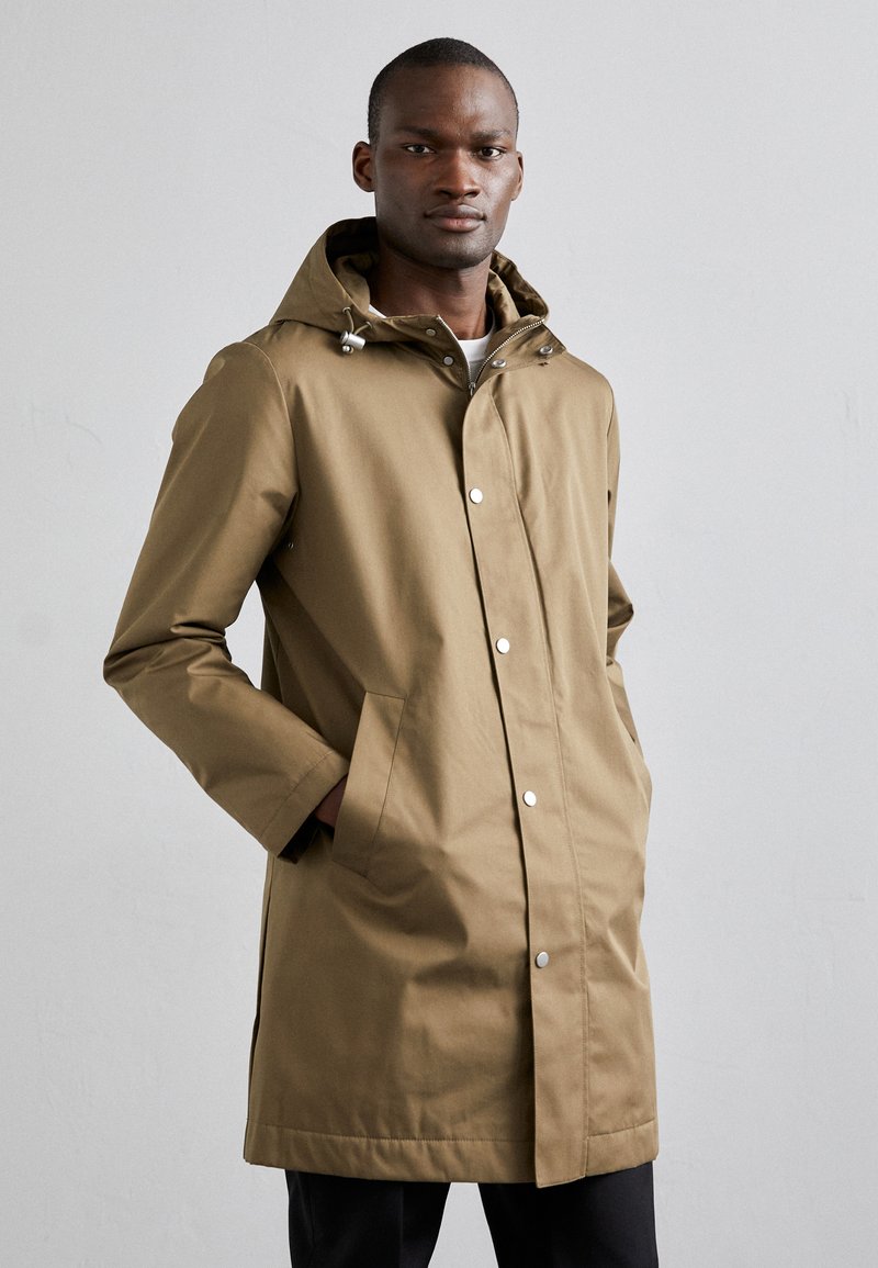 Cappotto impermeabile marrone con cappuccio, chiusura frontale a scatto e tasche laterali. Presenta una linea dritta e un tessuto testurizzato.