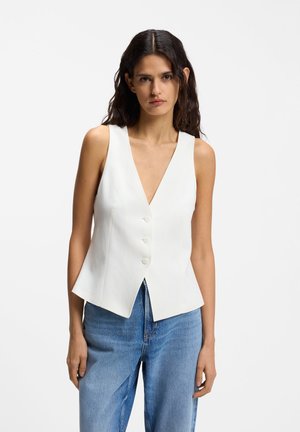 Kobieta z długimi, ciemnymi włosami, ubrana w białą, bezrękawnikową kamizelkę zapinaną na guziki oraz niebieskie dżinsy, stoi na tle jednolitego białego tła.