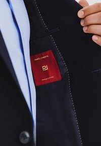 Granatowa marynarka o teksturowanym wykończeniu z czerwoną metką z napisem "PAKO LORENTE" oraz "PREMIUM SELECTION" w złotym kolorze. Widoczna jasnoniebieska koszula.