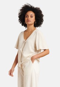 Beige Leinenoberteil mit V-Ausschnitt, kurzen Schlagärmeln und frontaler Knotendetail. Kombiniert mit hellen, gestreiften weit geschnittenen Hosen.