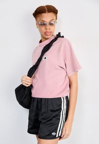 Pink cotton t-shirt with a small logo, paired with black satin shorts featuring white side stripes and an adidas emblem. Black bag slung over shoulder.